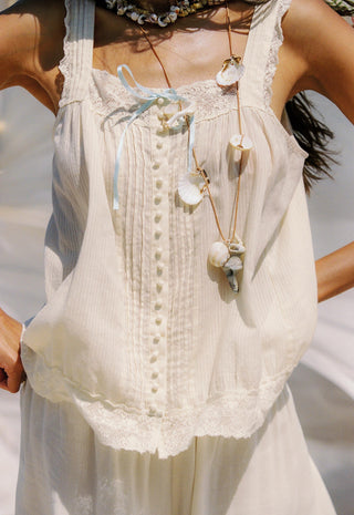 A woman wearing a cream lace-trimmed sleeveless top with a tie-front detail, layered with a shell necklace, and wearing a patterned headscarf against a blue ocean background.