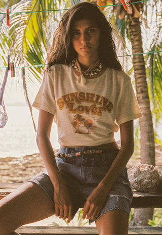 A woman sitting outdoors on a wooden bench wearing a cream graphic t-shirt that reads 'SUNGLING Lover' with a palm tree illustration, paired with denim shorts and a beaded belt. Palm trees and a beach are visible in the background.