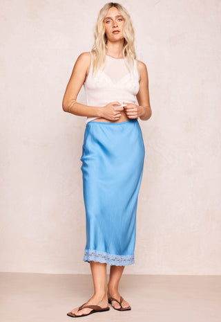 Woman with blonde beachy hair standing against a beige wall wearing a blue midi skirt with lace trim and a white tank top with brown leather sandals.