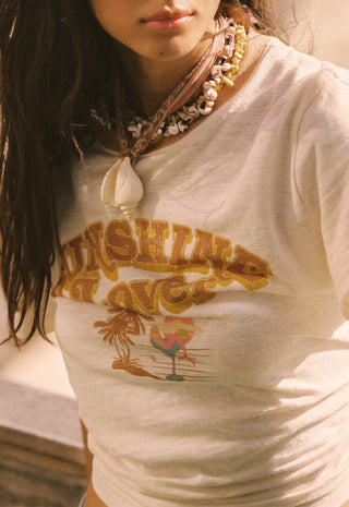 A close-up of a person wearing a white graphic t-shirt with a yellow 'Sunshine Ave.' text print and a tropical drink illustration, styled with layered shell necklaces.