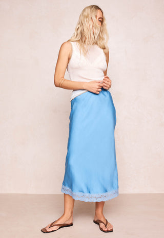 Woman with blonde beachy hair standing against a beige wall wearing a blue midi skirt with lace trim and a white tank top with brown leather sandals.