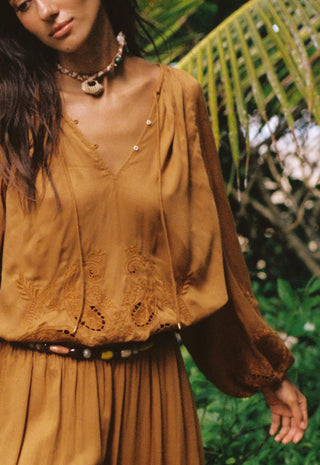A woman wearing a loose, earthy brown bohemian-style blouse with embroidered details and a matching pleated skirt, styled with a patterned necklace and belt, set against a lush green tropical background.