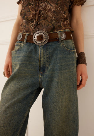 A close-up of a person wearing blue denim jeans with a brown leather belt featuring a large silver-toned western-style concho buckle, paired with a patterned brown top.