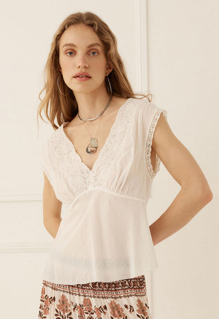 A woman wearing a white sleeveless lace-trimmed V-neck top with a gathered waist, paired with a brown and cream patterned skirt, standing against a white paneled wall.