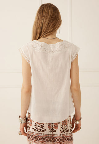 A woman viewed from behind wearing a cream-colored short-sleeve top with lace detailing around the neckline, paired with patterned pants.