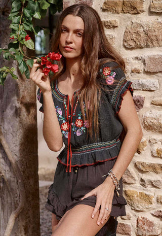 A woman wearing a dark gray embroidered blouse and matching shorts stands against a rustic stone wall, holding red flowers.
