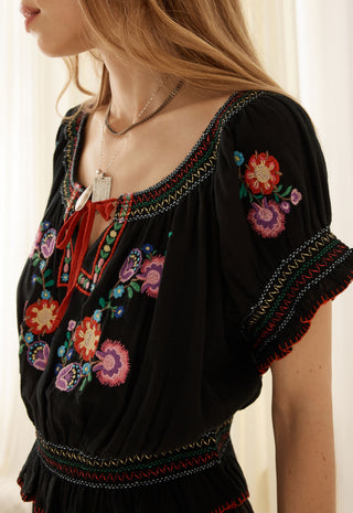 A close-up of a woman wearing a black bohemian-style blouse with colorful floral embroidery and a red tie-neck detail, paired with layered silver necklaces.