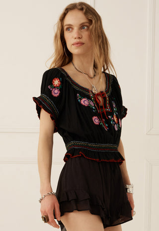 A woman wearing a black embroidered short-sleeve top and matching black ruffled shorts, standing against a white paneled wall.