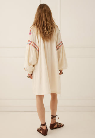 A woman viewed from behind wearing a cream-colored bohemian long-sleeve tunic dress with red and orange geometric embroidery on the sleeves, paired with brown strappy sandals, set against a white paneled wall.