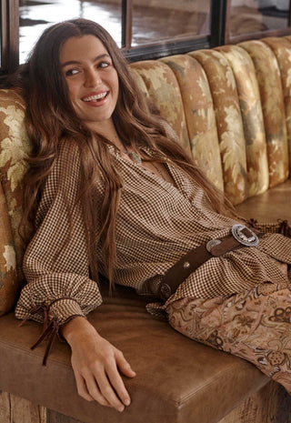 a-woman-with-long-brown-hair-reclines-on-a-brown-leather-bench-wearing-a-brown-and-beige-gingham-long-sleeve-top-with-a-matching-brown-belt-and-patterned-pants-set-against-a-rustic-background-with-a-patterned-gold-and-brown-velvet-sofa