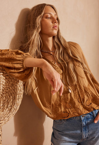 A woman wearing a tan eyelet lace blouse with puff sleeves and blue jeans, leaning against a textured beige wall next to a woven straw hat.