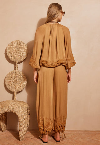 A woman stands with her back to the camera wearing a camel-brown two-piece set consisting of a loose long-sleeve top with lace trim and matching wide-leg pants with lace detailing at the hems. She is posed next to a woven rattan chair against a textured beige wall.