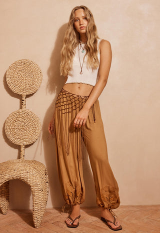 A woman stands indoors wearing loose tan harem-style pants with a black woven waist panel and drawstring, paired with a white sleeveless crop top and sandals. A woven chair is visible at left against a warm beige wall.