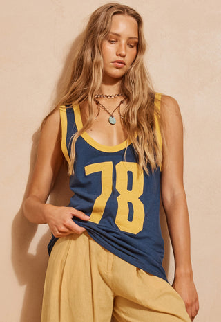 A woman wearing a navy blue tank top with yellow trim and a large yellow '78' on the front, paired with loose yellow pants, posed against a warm beige wall.