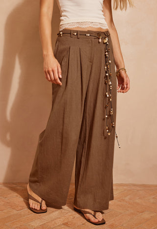 A model wearing high-waisted, wide-leg brown pants with a decorative beaded belt and a white lace-trimmed top, standing against a warm beige wall.