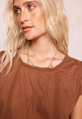 a-close-up-of-a-blonde-woman-wearing-a-brown-textured-top-and-layered-gold-necklaces-looking-off-camera-against-a-light-beige-background