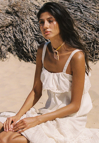 A woman sitting on a sandy beach wearing a white ruffled spaghetti-strap dress and a gold pendant necklace.