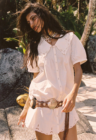 A woman wearing a white short-sleeve bohemian-style mini dress with eyelet lace details, a button-down front, and a wide brown woven belt with large shell-like accents, standing outdoors on a rocky tropical path.