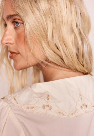 Close-up of a woman with blonde hair wearing a cream-colored blouse with a large embroidered collar.