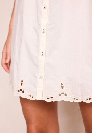 Close-up of a white eyelet button-front dress or tunic with a scalloped embroidered hem, shown on a model against a plain light beige background.