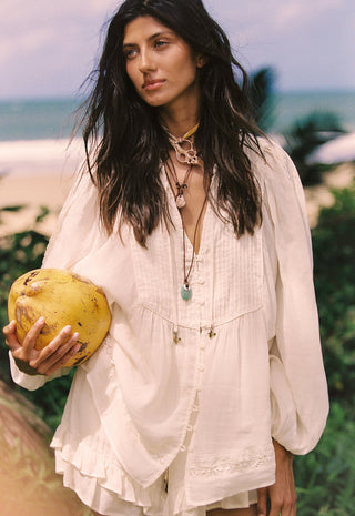 A woman on a beach wearing a loose white bohemian-style blouse and holding a yellow coconut