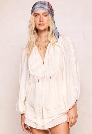 A woman wearing a cream-colored, long-sleeved button-up blouse with pleated details and a matching tiered skirt, styled with a blue patterned headscarf against a textured beige background.