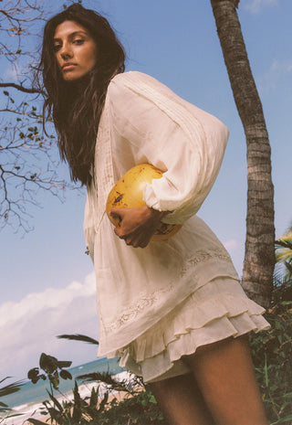 A woman wearing a loose white long-sleeve mini dress with lace trim stands outdoors near a palm tree, holding a yellow fruit against a blue sky background.