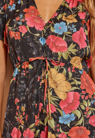 A close up shot of a woman modelling a short-sleeve floral mini dress with a lace hem, standing against a textured beige wall.