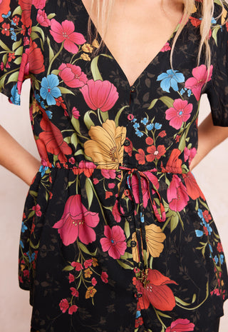 A close-up of a black short-sleeve V-neck dress with a vibrant floral print featuring pink, red, blue, and yellow flowers with green leaves. The dress has a drawstring waist and button details down the front.