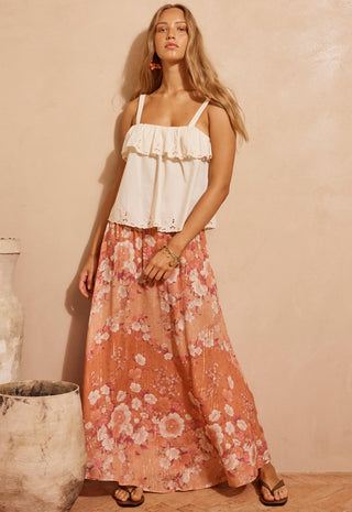 A woman modeling a white ruffled camisole top paired with a long peach-pink floral maxi skirt, standing against a warm beige textured wall.
