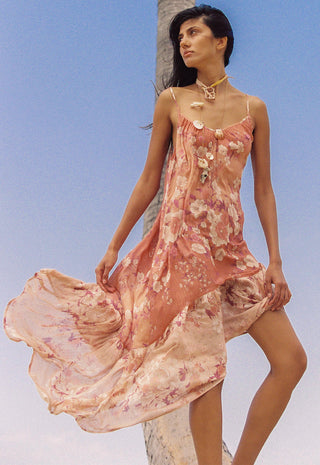 A woman wearing a flowing floral slip dress in soft pink and peach tones, posed outdoors against a clear blue sky with a palm tree visible behind her.