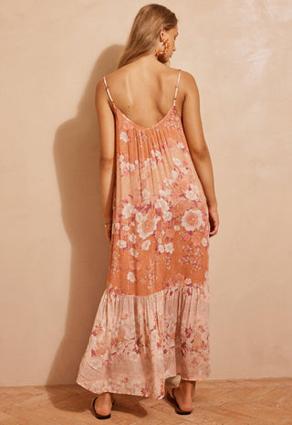 A woman wearing a peach and pink floral maxi dress with spaghetti straps, shown from the back against a textured beige wall.