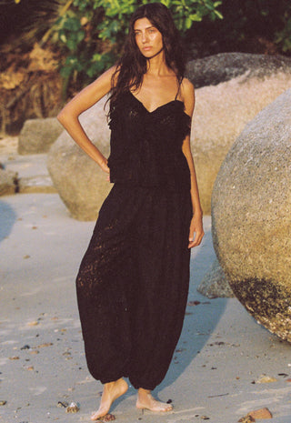 A woman stands barefoot on a sandy beach with large rocks and greenery in the background, wearing a black lace camisole and matching black harem-style pants.