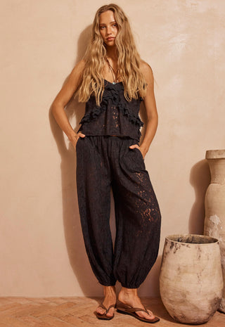 A woman modeling a black lace sleeveless top and matching black lace harem pants, standing in a warm beige studio setting with decorative ceramic pots.