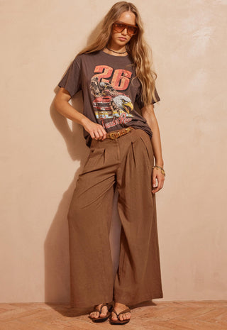 A woman modeling a dark brown graphic t-shirt and matching brown wide-leg pleated trousers, styled with a brown belt, sunglasses, and sandals against a textured beige wall.