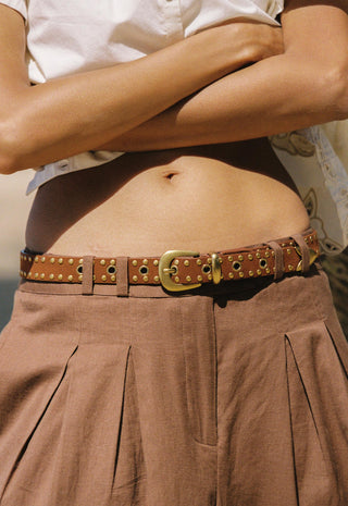 a-close-up-of-a-person-wearing-a-brown-studded-leather-belt-with-a-gold-tone-buckle-paired-with-a-white-top-and-pleated-brown-skirt