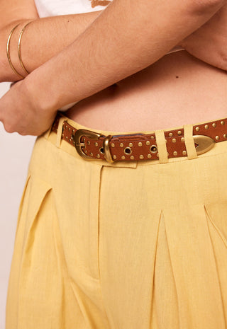 a-close-up-of-a-model-wearing-a-brown-leather-belt-with-brass-studs-and-a-gold-tone-buckle-paired-with-pleated-yellow-trousers