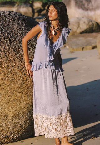 A woman stands outdoors on a sandy beach next to a large rock, wearing a light lavender sleeveless maxi dress with ruffled shoulders and a cream lace hem.