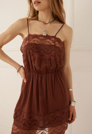 A woman wearing a brown lace-trimmed spaghetti-strap camisole dress with a gathered empire waist, shown from the neck down against a light cream paneled wall.