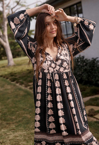 A woman wearing a black bohemian-style maxi dress with a pink floral print, standing outdoors in a grassy yard.