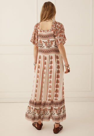 A woman viewed from behind wearing a bohemian-style maxi dress with brown and beige patterns, standing against a white paneled wall.