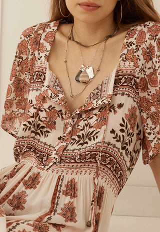 A close-up of a woman wearing a bohemian-style cream dress with rust-orange and dark brown floral patterns, layered with silver necklaces.