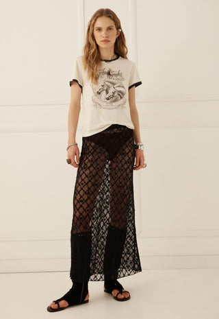 A model stands against a white paneled wall wearing a cream graphic t-shirt with black trim, sheer black patterned maxi pants, and black sandals.