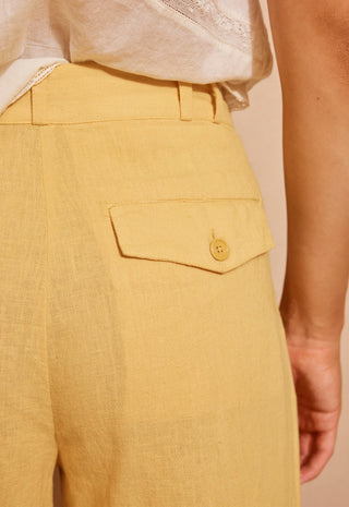 Close-up of a person wearing mustard yellow linen trousers with a buttoned flap pocket and a white top, against a plain beige background.