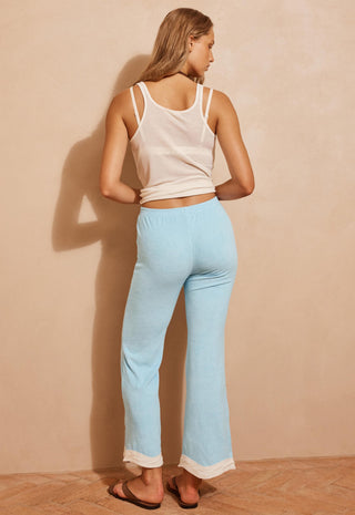 A woman stands with her back to the camera wearing light blue flared lounge pants with a white hem, paired with a white tank top and brown sandals, against a warm beige wall.