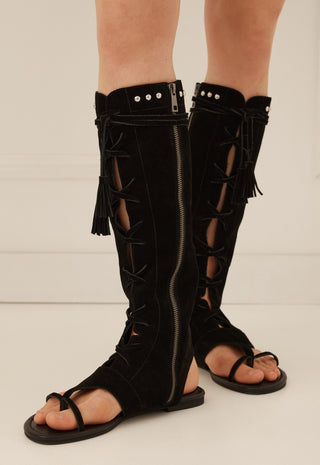 Black suede knee-high strappy sandals with side zippers, cutout details, and fringe, shown on a model's feet against a light neutral background.